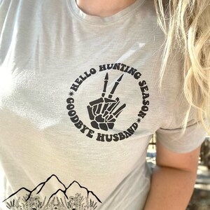 May include: A light gray t-shirt with a black graphic of a skeleton hand making a peace sign. The graphic is surrounded by a circle with the text "Hello Hunting Season Goodbye Husband".