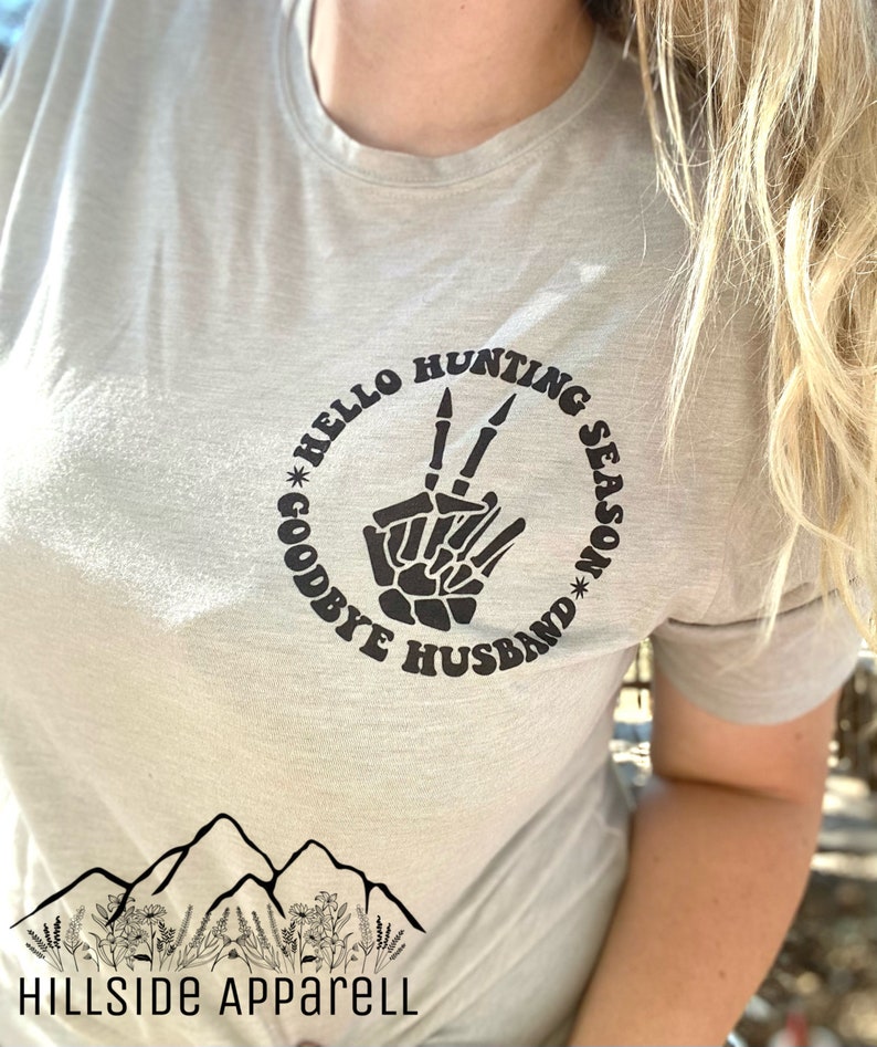 May include: A light gray t-shirt with a black graphic of a skeleton hand making a peace sign. The graphic is surrounded by a circle with the text "Hello Hunting Season Goodbye Husband".