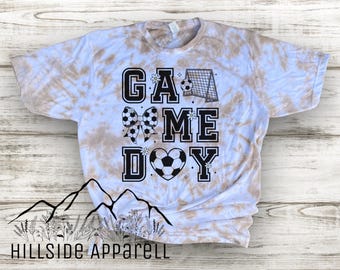May include: Tie-dye t-shirt with the words "GAME DAY" in black block letters. The design includes soccer balls, a goal, and a soccer ball heart. The shirt has a light brown and white tie-dye pattern.