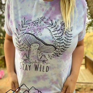 K&ouml;nnte beinhalten: Ein Batik-T-Shirt in Lila- und Blaut&ouml;nen. Das Shirt zeigt eine schwarz-wei&szlig;e Illustration eines Pilzes mit Bl&auml;ttern und dem Text "Stay Wild".