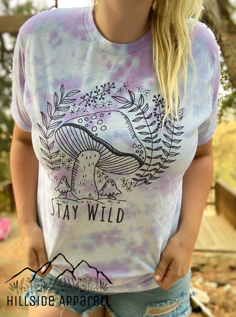 K&ouml;nnte beinhalten: Ein Batik-T-Shirt in Lila- und Blaut&ouml;nen. Das Shirt zeigt eine schwarz-wei&szlig;e Illustration eines Pilzes mit Bl&auml;ttern und dem Text "Stay Wild".