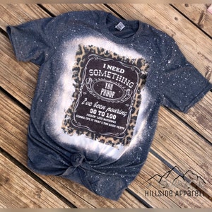 May include: A dark gray t-shirt with a bleached design featuring a leopard print background and a white graphic that reads "I need something you proof I've been pouring 90 to 100 feelin' like nothing gonna cut it that's the hard truth".