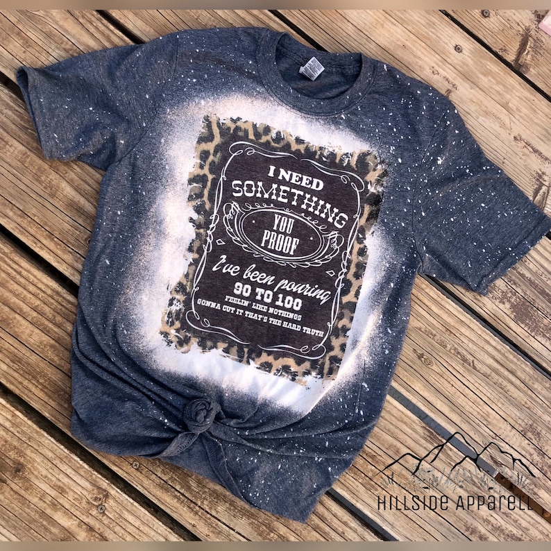 May include: A dark gray t-shirt with a bleached design featuring a leopard print background and a white graphic that reads "I need something you proof I've been pouring 90 to 100 feelin' like nothing gonna cut it that's the hard truth".