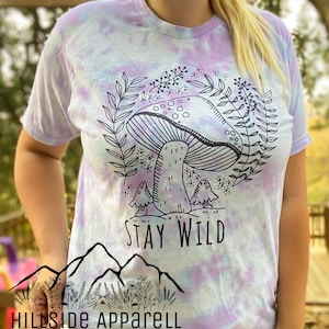 K&ouml;nnte beinhalten: Ein Batik-T-Shirt mit einem Pilz-Motiv und dem Text "Stay Wild". Das Shirt ist hellblau, lila und wei&szlig;.