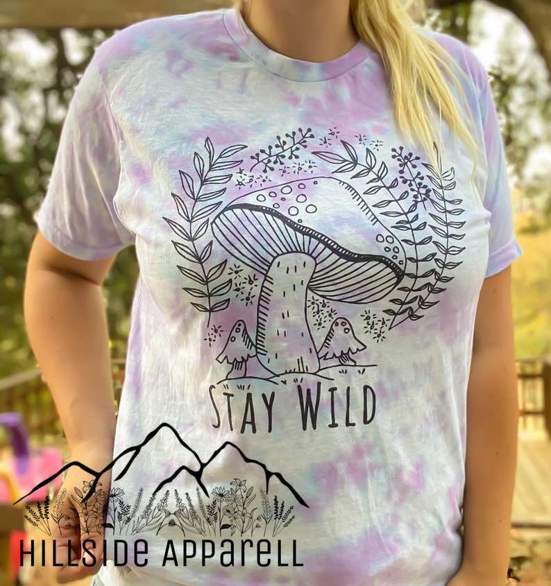 K&ouml;nnte beinhalten: Ein Batik-T-Shirt mit einem Pilz-Motiv und dem Text "Stay Wild". Das Shirt ist hellblau, lila und wei&szlig;.