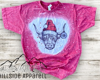 May include: Red and white bleached t-shirt with a graphic of a highland cow wearing a Santa hat and holding an ornament. The shirt has a distressed look with white speckles.