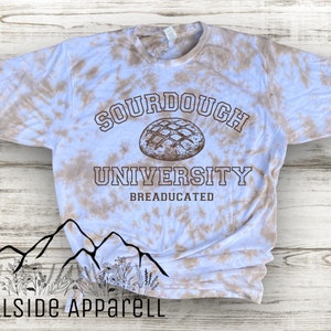 May include: A light blue tie-dye t-shirt with the text "Sourdough University" and "BREADUCATED" in brown. A drawing of a sourdough bread loaf is above the text.