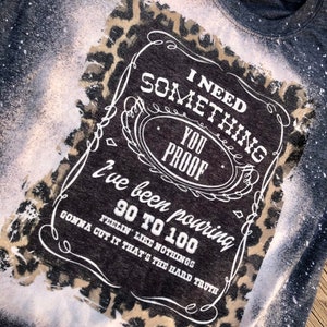 May include: A black t-shirt with a white bleach design of a whiskey bottle label. The label says "I need something you proof I've been pouring 90 to 100. Feelin' like nothings gonna cut it that's the hard truth." The design is surrounded by a leopard print.