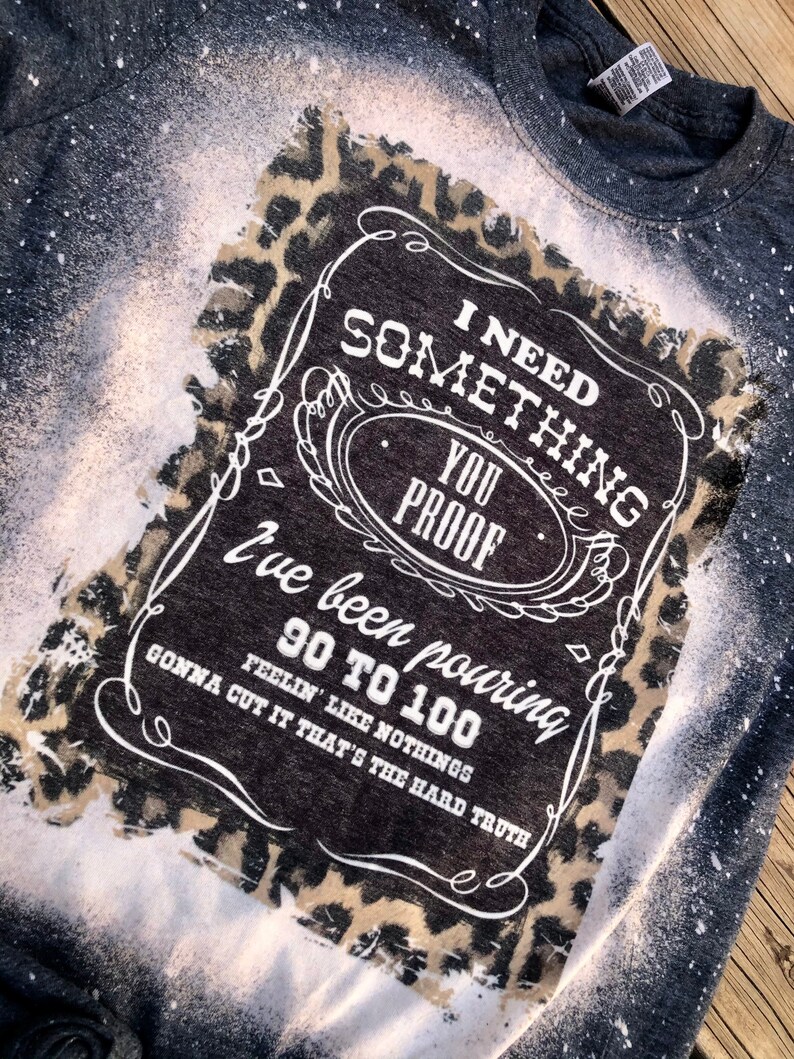 May include: A black t-shirt with a white bleach design of a whiskey bottle label. The label says "I need something you proof I've been pouring 90 to 100. Feelin' like nothings gonna cut it that's the hard truth." The design is surrounded by a leopard print.