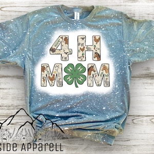 May include: A light blue t-shirt with a white splattered design. The shirt features the text "4-H MOM" with animal illustrations inside the letters and a green four-leaf clover. The brand "HILLSIDE APPARELL" is printed at the bottom.