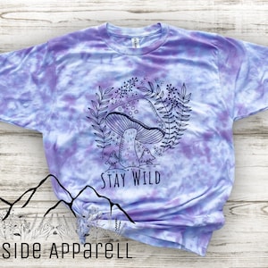 K&ouml;nnte beinhalten: Lilafarbenes T-Shirt im Tie-Dye-Stil mit einer schwarz-wei&szlig;en Illustration eines Pilzes, Bl&auml;ttern und V&ouml;geln. Der Text "Stay Wild" ist unter der Illustration aufgedruckt.