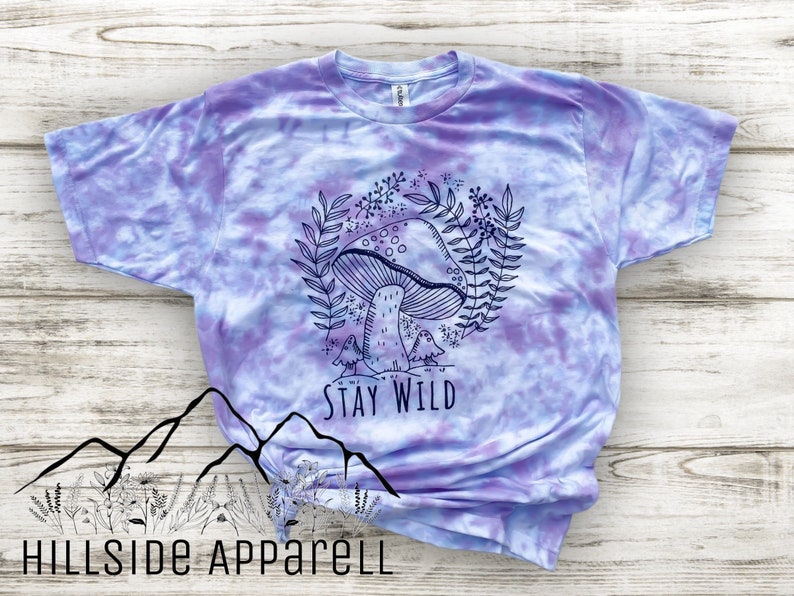 K&ouml;nnte beinhalten: Lilafarbenes T-Shirt im Tie-Dye-Stil mit einer schwarz-wei&szlig;en Illustration eines Pilzes, Bl&auml;ttern und V&ouml;geln. Der Text "Stay Wild" ist unter der Illustration aufgedruckt.