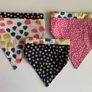 May include: Three dog bandanas with different patterns. One bandana is white with pink, orange, and blue abstract shapes. Another bandana is pink with white polka dots. The third bandana is navy blue with a small floral pattern in pink, yellow, and blue.