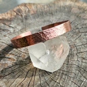 May include: A hammered copper cuff bracelet with a smooth, polished finish. The bracelet is displayed on a wooden surface with a clear quartz crystal.