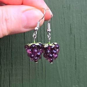 May include: A pair of grape cluster earrings. Each earring features a cluster of purple beads, silver-coloured hooks, and green accents. The earrings are held up by a hand against a green backdrop.