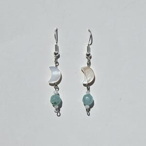 May include: Dangle earrings with a crescent moon, small white bead, and a larger, round, light blue bead. The earrings have silver-coloured hooks and are shown against a white backdrop. The earrings are a jewellery item.