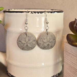 May include: A pair of grey sand dollar earrings. Each earring has a detailed sand dollar design with a silver hook. The earrings are shown against a white mug.