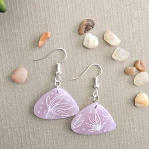 May include: Lavender-coloured, triangular earrings with a white floral design. The earrings hang from silver-coloured hooks. Small white dots accent the floral pattern. The earrings are displayed on a neutral surface with small decorative stones.