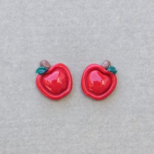 May include: A pair of stud earrings shaped like glossy red apples. Each apple features a smooth, rounded red body with a small brown stem and a single green leaf attached at the top. The surface of the apples has a shiny, reflective finish.
