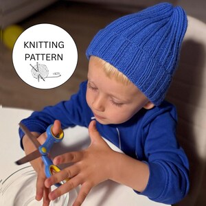 Könnte beinhalten: Eine blaue Strickmütze. Die Mütze wird von einer Person getragen, die einen blauen Pullover trägt. Das Bild enthält einen weißen Kreis mit den Worten "KNITTING PATTERN" und eine Wollknäuel-Illustration. Die Person hält eine blaue und gelbe Schere.