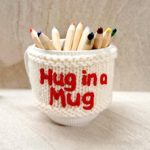 May include: A white ceramic mug with a knitted cozy that reads "Hug in a Mug" in red letters. The mug is filled with colored pencils, each with a different colored tip.