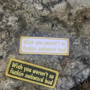 May include: Two embroidered patches with gold thread. The top patch is white with gold thread and says "Wish you weren't so fuckin awkward bud". The bottom patch is dark green with gold thread and says "Wish you weren't so fuckin awkward bud".