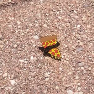 May include: A pizza slice enamel pin with a brown crust, red sauce, yellow cheese, and black text that reads "Pizza Slut". The pin is set against a textured, reddish-brown surface.