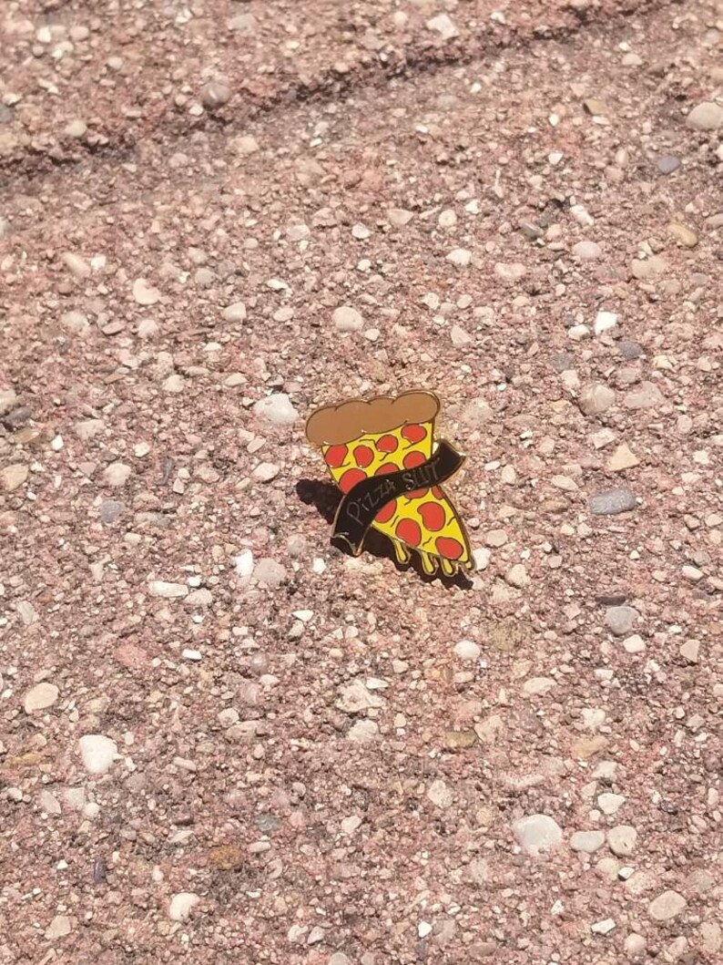 May include: A pizza slice enamel pin with a brown crust, red sauce, yellow cheese, and black text that reads "Pizza Slut". The pin is set against a textured, reddish-brown surface.
