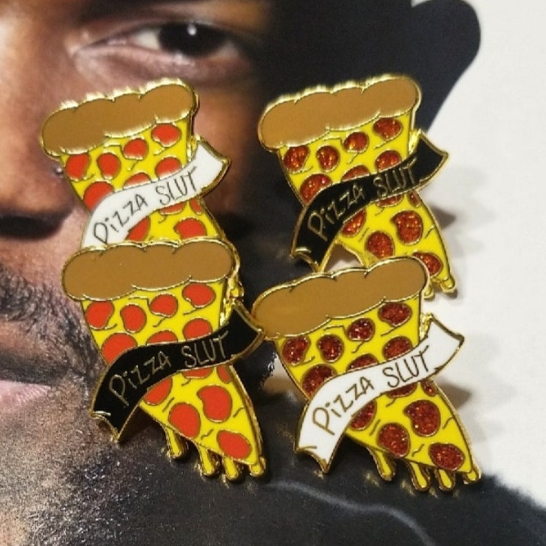 May include: Four pizza slice enamel pins. Each pin features a yellow pizza slice with red pepperoni and a brown crust. A banner across each slice reads "Pizza Slut" in black or white text. The pins have a gold outline.