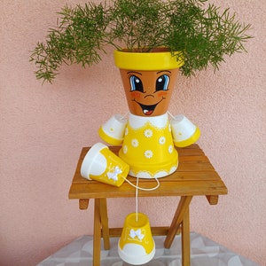 Könnte beinhalten: Ein gelber und weißer bemalter Blumentopf mit einem lächelnden Gesicht und einem Gänseblümchenmuster. Der Topf hat drei kleinere Töpfe, die daran hängen. Der Topf steht auf einem klappbaren Holztisch.