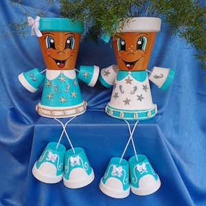 Puede incluir: Dos macetas de terracota pintadas con caras sonrientes, que llevan trajes de marinero azul y blanco y zapatos blancos con ribetes azules. Las macetas están decoradas con conchas marinas, estrellas de mar e hipocampos. Las macetas están sentadas sobre una superficie azul.