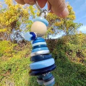 Könnte beinhalten: Handgefertigter Schlüsselanhänger mit einer Knopfpuppe. Die Puppe besteht aus blauen Knöpfen in verschiedenen Farbtönen, einem Holzkopf und einem silberfarbenen Schlüsselring. Der Hintergrund zeigt grünes und gelbes Laub. Ein einzigartiges Accessoire.
