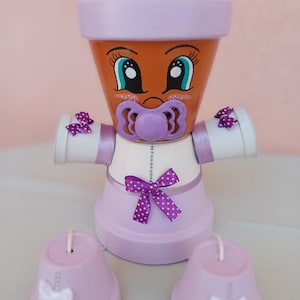 Può includere: Una figura decorativa realizzata con vasi in terracotta, caratterizzata da un volto dipinto con grandi occhi e un ciuccio viola. La figura indossa un abito bianco e viola chiaro decorato con fiocchi viola a pois e due vasi più piccoli alla base.