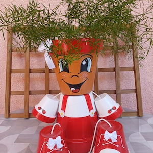 Op de afbeelding: Een grillige, handgemaakte terracotta bloempot in de vorm van een lachend karakter, met een groene plant die uit de bovenkant ontspruit. De pot heeft geschilderde details, waaronder ogen, een mond en rood-witte kleding. Twee bijpassende rood-witte schoenvormige potten staan ervoor.
