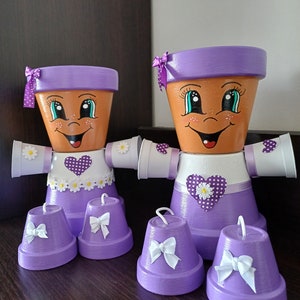Può includere: Due vasi di terracotta dipinti di viola con volti, cuori a pois bianchi e dettagli di margherite bianche. I vasi sono decorati con fiocchi viola e hanno vasi più piccoli di colore viola con fiocchi bianchi attaccati alla parte inferiore.