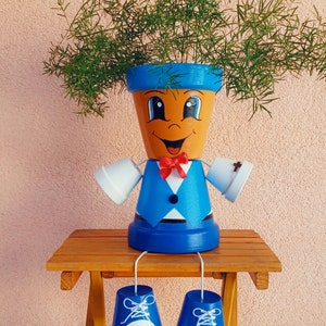 Puede incluir: Una maceta de terracota pintada a mano con forma de persona, con una cara sonriente, un traje azul y una pajarita roja. La maceta tiene brazos blancos y zapatos azules y blancos. Las plantas verdes brotan de la parte superior.