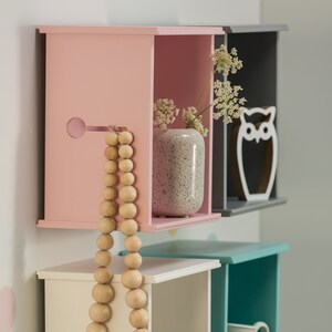 May include: Four square wooden shelves in pink, grey, white, and teal with decorative items on each shelf. The top shelf has a small vase with white flowers and a white owl figurine. The second shelf has a wooden bead necklace. The third shelf has a small grey elephant figurine. The bottom shelf has a white jug.