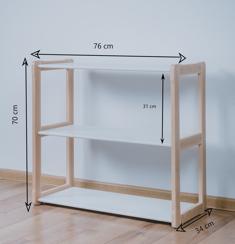 Puede incluir: Estanter&iacute;a de madera de tres niveles con estantes blancos. La estanter&iacute;a mide 76 cm de ancho, 70 cm de alto y 34 cm de profundidad. El marco es de madera clara y los estantes son blancos.