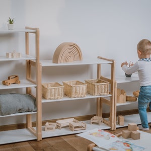 May include: A white bookshelf with wooden accents, filled with wooden toys, baskets, and a rainbow arch. A young child is standing in front of the shelf, reaching for a toy.