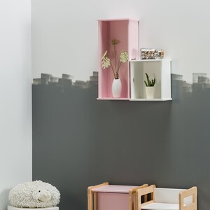 May include: A pink and white children's storage bench with a white fluffy toy on top. The bench has a drawer and a seat. The bench is in front of a wall with a pink and white shelf with a vase of flowers and a small plant.
