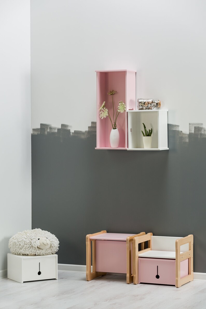May include: A pink and white children's storage bench with a white fluffy toy on top. The bench has a drawer and a seat. The bench is in front of a wall with a pink and white shelf with a vase of flowers and a small plant.