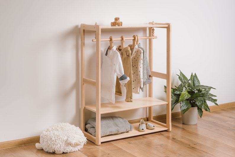 May include: A wooden clothes rack with two shelves and a hanging bar. The rack is made of natural wood and has a simple design. There are clothes hanging on the bar, including a white shirt, a pair of pants, and a patterned shirt.