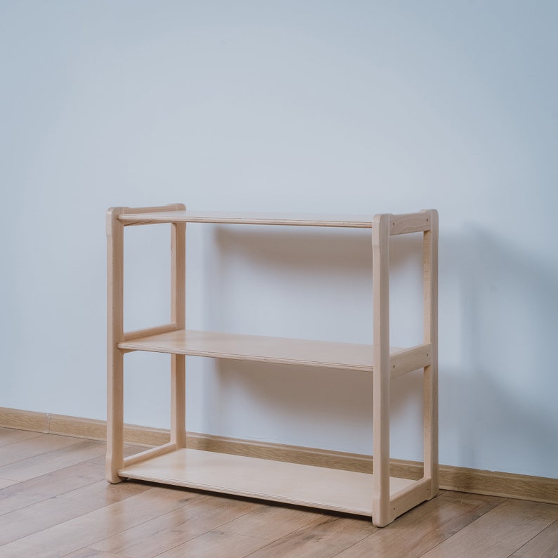 May include: A light wood shelf unit with three shelves. The shelf unit is standing on a light wood floor.