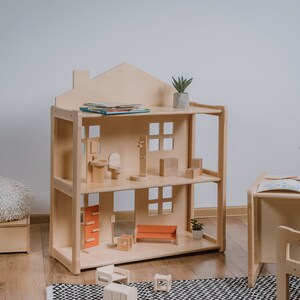 May include: A wooden dollhouse with two levels and a roof. The dollhouse is filled with miniature furniture, including a bed, a couch, a toilet, and a sink. There are also some books on the top shelf. The dollhouse is sitting on a wooden floor.
