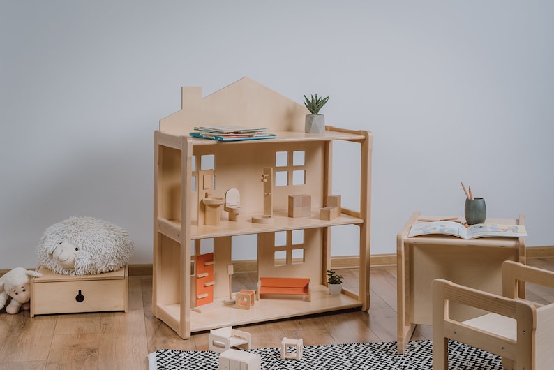 May include: A wooden dollhouse with two levels and a roof. The dollhouse is filled with miniature furniture, including a bed, a couch, a toilet, and a sink. There are also some books on the top shelf. The dollhouse is sitting on a wooden floor.