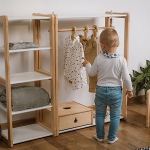 May include: A wooden children's clothing rack with shelves and a hanging rod. A young child is standing in front of the rack, looking at the clothes.