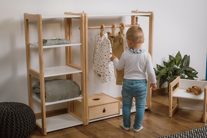 May include: A wooden children's clothing rack with shelves and a hanging rod. A young child is standing in front of the rack, looking at the clothes.
