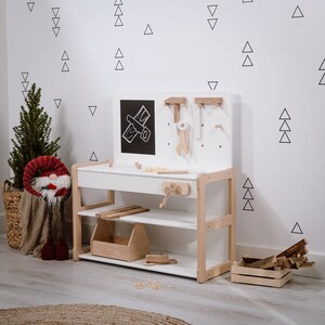 Puede incluir: Un banco de trabajo para niños de madera y blanco con un panel de pizarra, estantes y un estante para herramientas. El banco de trabajo tiene un diseño en blanco y negro de un avión en el panel de pizarra. El banco de trabajo está frente a una pared blanca con calcomanías triangulares negras.