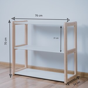 May include: White shelf unit with three shelves and a natural wood frame. The shelf unit is 76 cm wide, 70 cm tall, and 34 cm deep.