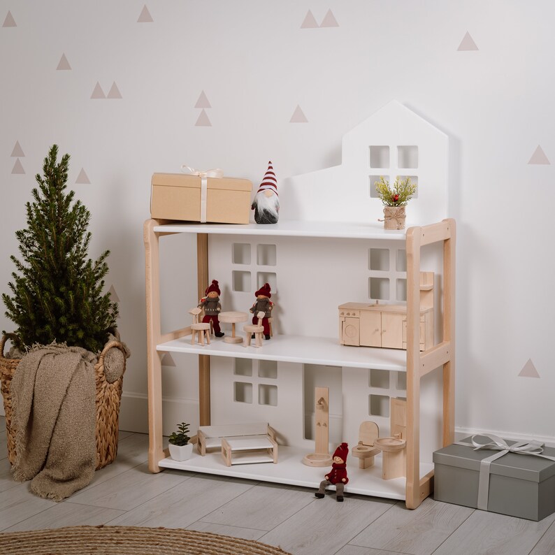 Puede incluir: Una casa de mu&ntilde;ecas de madera blanca de tres niveles, con muebles y accesorios en miniatura. La casa de mu&ntilde;ecas tiene una parte superior en forma de casa con ventanas. La escena incluye un peque&ntilde;o &aacute;rbol de Navidad, una caja de regalo y gnomos decorativos.